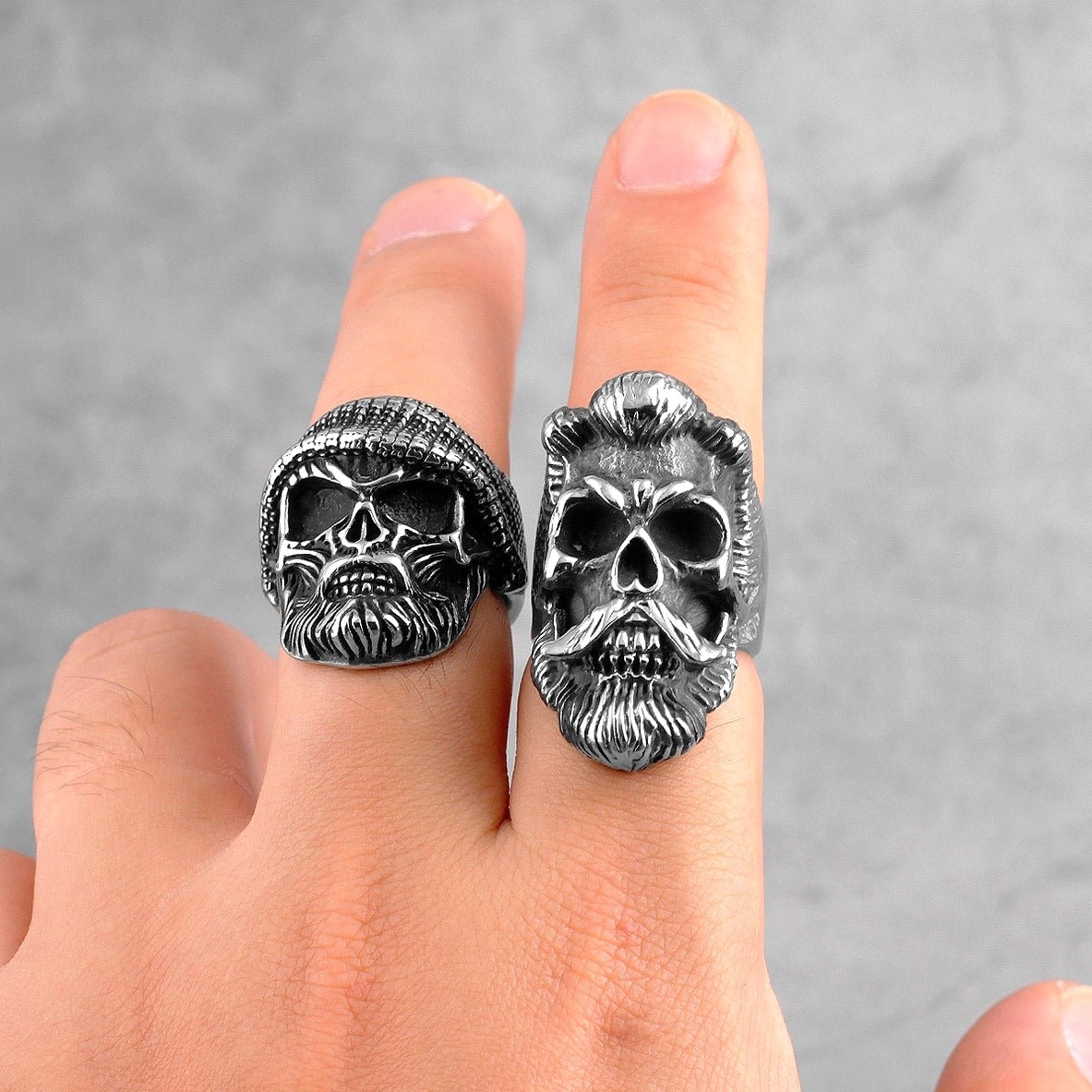 Two bearded skull gothic rings worn on human fingers one with knit beanie detail grey background