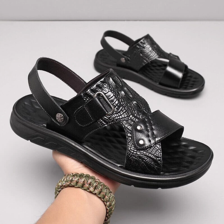 Black leather sandals held in hand showcasing crocodile embossed texture and genuine leather craftsmanship