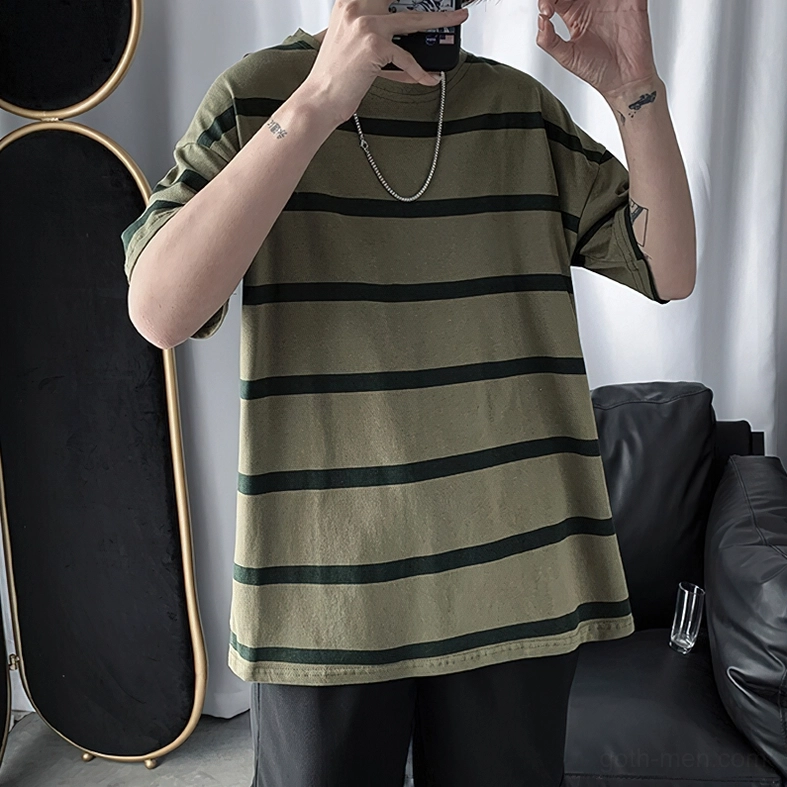 Male model wearing oversized striped gothic t-shirt olive base with dark green stripes, styled with silver chain necklace, front upper-body indoor home setting