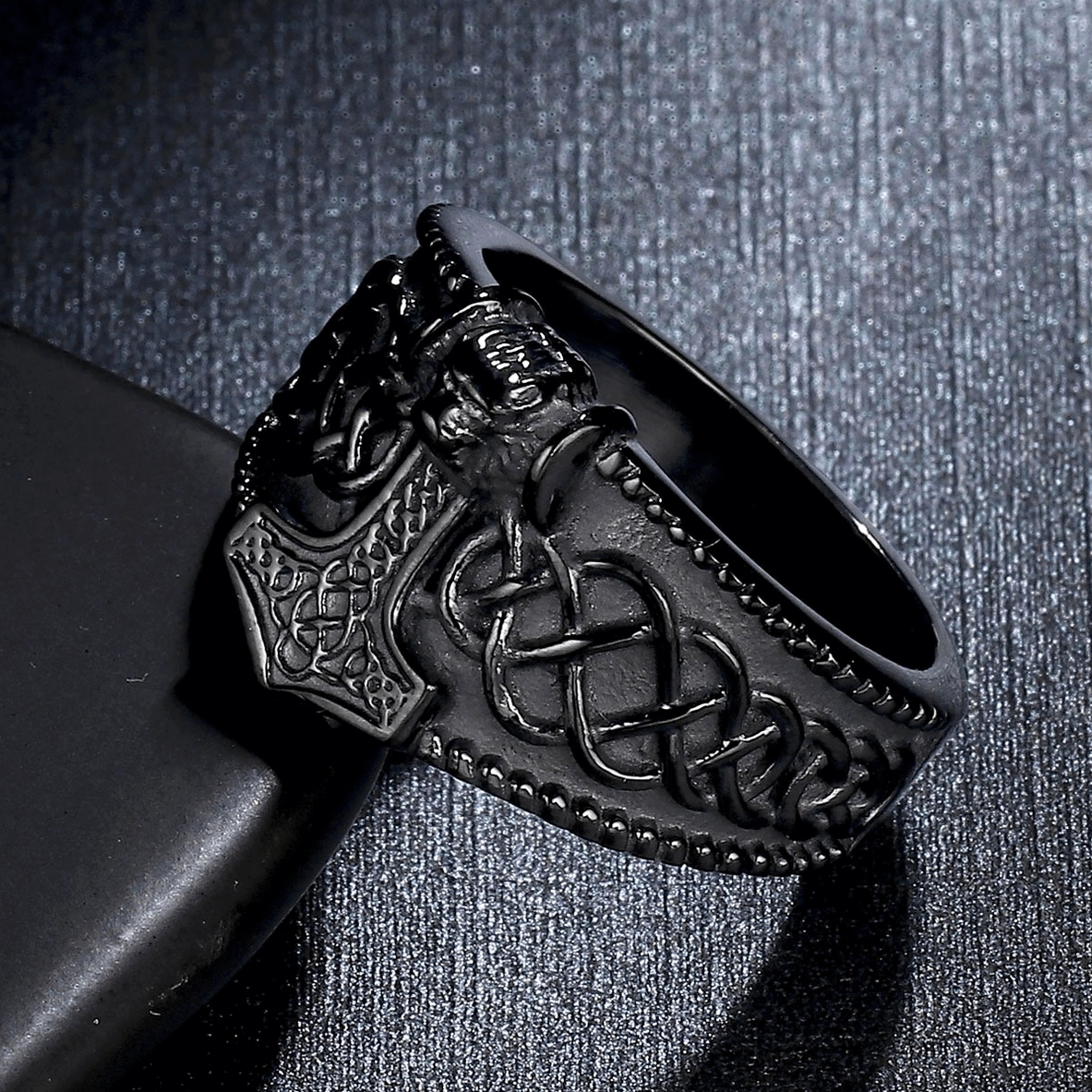 Side oblique view of matte black gothic men's ring showing Mjolnir, skull, and Celtic knot depth on dark backdrop